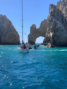 Two boats navigating through turquoise waters near a large rock formation that features a natural arch. The sky is clear and blue, with sunlight reflecting off the water. Los Cabos San Lucas, México 2019.