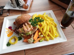 A chicken burger on a sesame seed bun served on a white rectangular plate with potato chips, steamed vegetables, and colorful sauces, alongside a glass of dark soda on a wooden table.