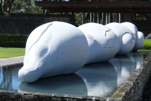 A series of large, smooth, white, rounded stone sculptures forming a curved line, partially submerged in a shallow water feature, with green lawn, stone wall, and wooden structure in the background.
