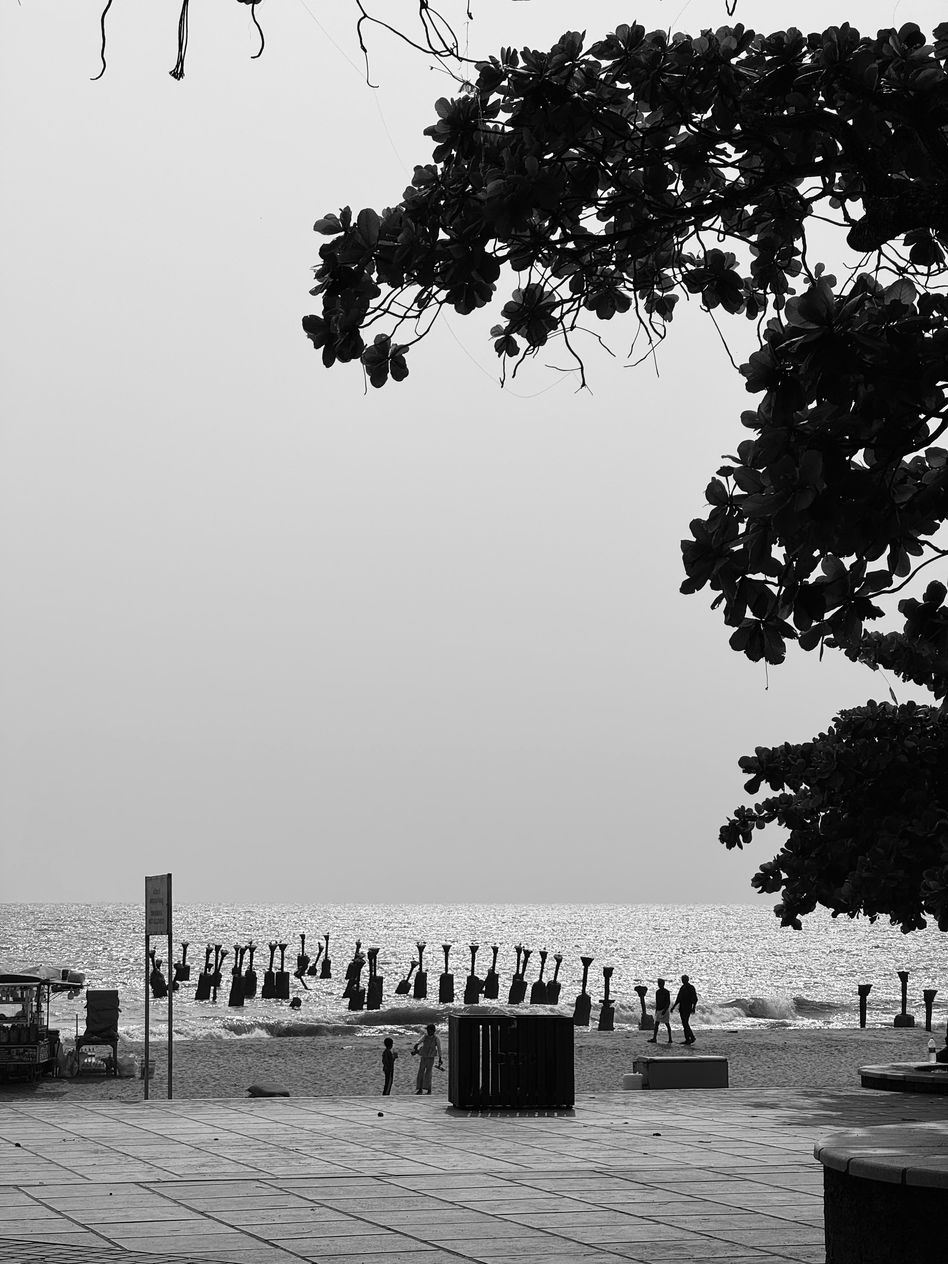 A black-and-white photograph capturing a beach scene. In the foreground, there are silhouettes of a few people standing and walking on the shore, near some broken pier poles that rise from the sand into the background.