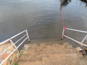 Weathered concrete steps lead down to the shore of Lake Victoria, with white railings on both sides and a red rope tied to one side extending into the water.