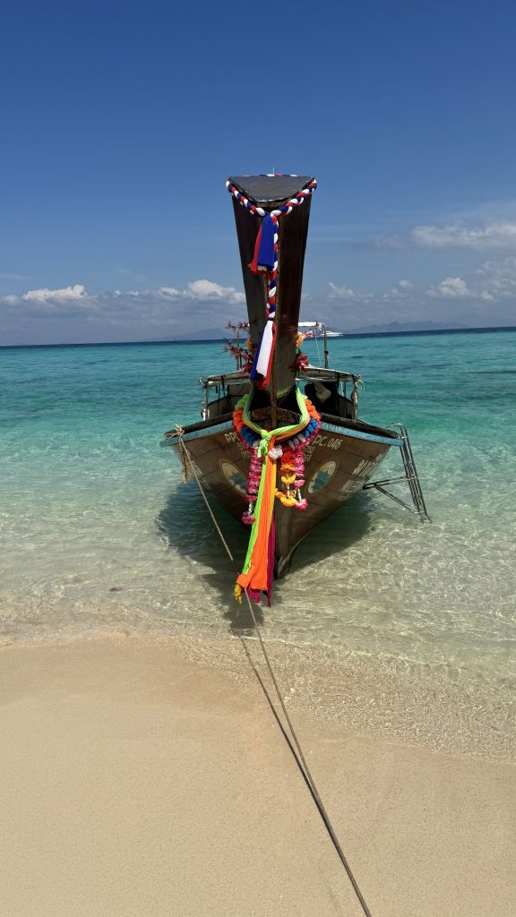 A wooden longtail boat sits in clear turquoise water by a sandy beach. It has colorful garlands on the front, a rope tied to the shore, and the bright blue sky reflects in the water.