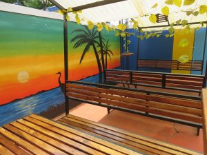 An outdoor seating area features several wooden slat benches and tables under a translucent roof. A wall mural depicts a sunset over water with palm trees and a bird silhouette. Artificial green vines hang from the roof structure.
