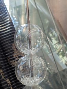 A close-up photograph of a decorative object consisting of two clear glass spheres stacked vertically. Each sphere contains small pieces of white crystalline material at the bottom. A metallic rod is visible passing through the center of the spheres.
