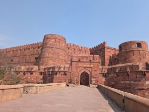 Red sandstone fort with high walls, circular bastions, and an arched gateway, approached by a stone bridge.