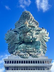 A large stone statue of Lord Vishnu seated on Garuda with outstretched wings, richly carved and set on a base against a bright sky with clouds.