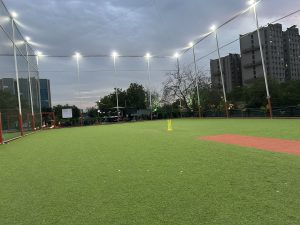 A wide-angle view of a professional outdoor cricket turf under stadium floodlights at dusk, featuring a green artificial grass surface, a red pitch, yellow wickets, and tall safety netting with urban buildings in the background.