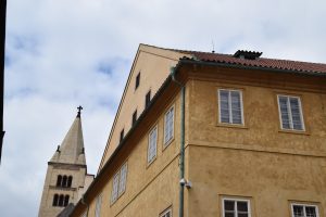 Low angle view of historic yellow buildings in Prague. Features a stone church tower in the background, red-tiled roofs, and a cloudy sky.