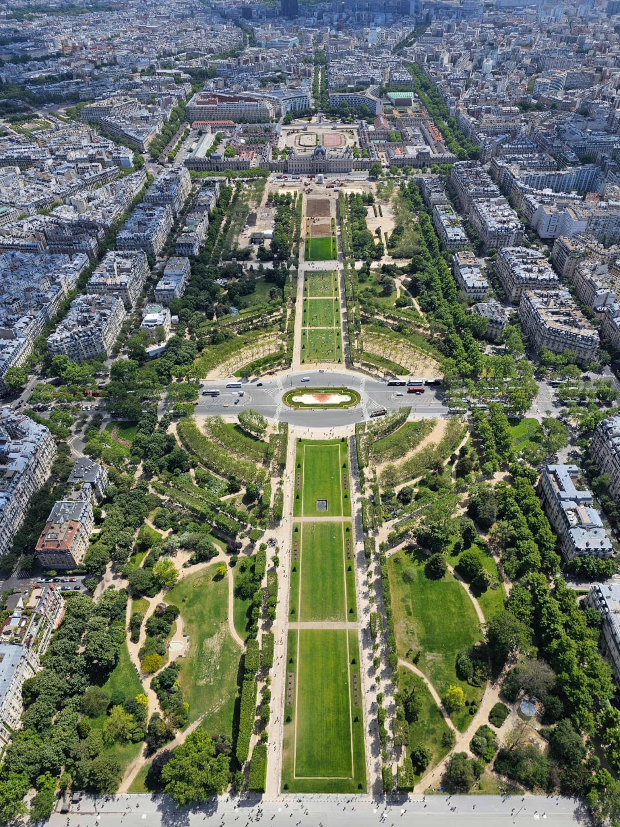 This image shows a perfectly symmetrical aerial view of a large urban park stretching through the middle of a dense city. The park features long, rectangular lawns, tree-lined pathways, and carefully designed gardens arranged in a geometric pattern.
