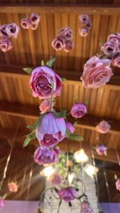 A close-up view of pink artificial roses hanging from a string against a wooden ceiling. The roses have vibrant colors and green leaves, creating a whimsical atmosphere. In the background, there are hints of lighting fixtures and decorative elements, contributing to the overall aesthetic.