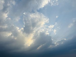 A view looking up at a blue sky filled with textured, dramatic cumulus clouds. Sunlight breaks through gaps in the clouds, creating visible sunbeams that radiate across the sky.