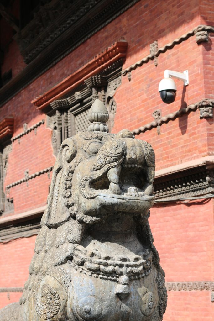 A close-up of a stone lion statue, also known as a guardian lion or “shishi,” displaying intricate carvings and ornate details on its mane and body. The statue stands in front of a textured red brick wall, which features traditional architectural elements, including wooden carvings and a modern security camera mounted on the wall. The sunlight casts shadows, emphasizing the depth of the statue’s features.