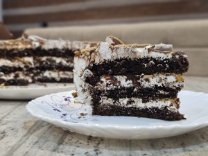 A close-up of a decadent slice of multi-layered chocolate cake with white frosting and caramel drizzle on a decorative plate.
