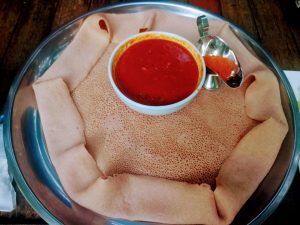A large, circular piece of injera bread with a porous texture is spread across a metal tray, with additional pieces rolled along the edges. A white bowl containing a red sauce is placed in the center. A small metal boat-shaped container with red spice powder is positioned to the right of the bowl. The tray sits on a wooden surface.