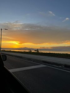 A scenic view of a sunset over the ocean, with vibrant orange and yellow hues illuminating the sky. The horizon showcases distant mountains, and a paved road is visible in the foreground with a traffic light and foliage along the roadside.