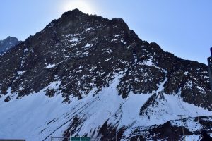 A rugged mountain peak covered in snow, with a bright sun partially hidden behind the summit. The lower slopes show a mix of rocky terrain and patches of snow, while a clear blue sky fills the background. Road signs are visible at the base, suggesting a nearby roadway.