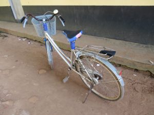 A silver bicycle with a front basket parked on a dirt surface beside a building wall.