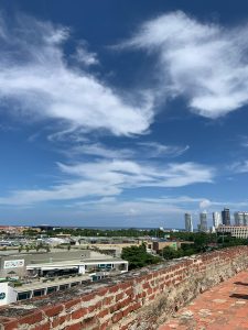 A wide city view from atop an old brick fortress wall with modern skyscrapers and the ocean visible in the distance
