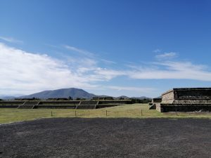 Wide landscape of the Temple of Quetzalcoatl with volcanic soil, ancient ruins, green hills, and a distant mountain under a blue sky.