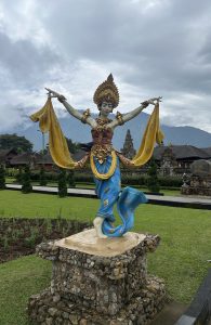 A colorful statue of a traditional Balinese dancer in a graceful pose, with arms extended and flowing yellow scarves. The figure is adorned with an ornate headdress and detailed costume, standing on a stone pedestal within a lush garden, with temple buildings and cloudy skies in the background.