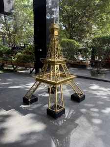 A golden miniature replica of the Eiffel Tower stands in a landscaped area with lush green plants in the background.