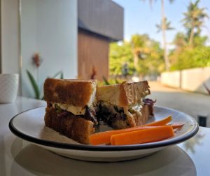 A thick-cut, toasted sandwich sliced in half and served on a white plate with carrot sticks, set against a bright outdoor background.
