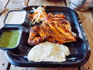 A black rectangular plate features a piece of grilled chicken, characterized by a golden-brown and charred exterior.
