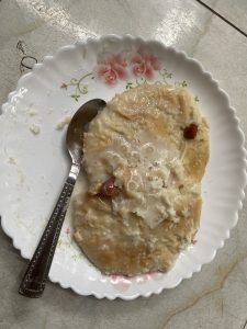 A white plate with floral patterns holds a portion of milk-soaked malpua, garnished with nuts. A spoon rests on the plate. The scene is casual and inviting.
