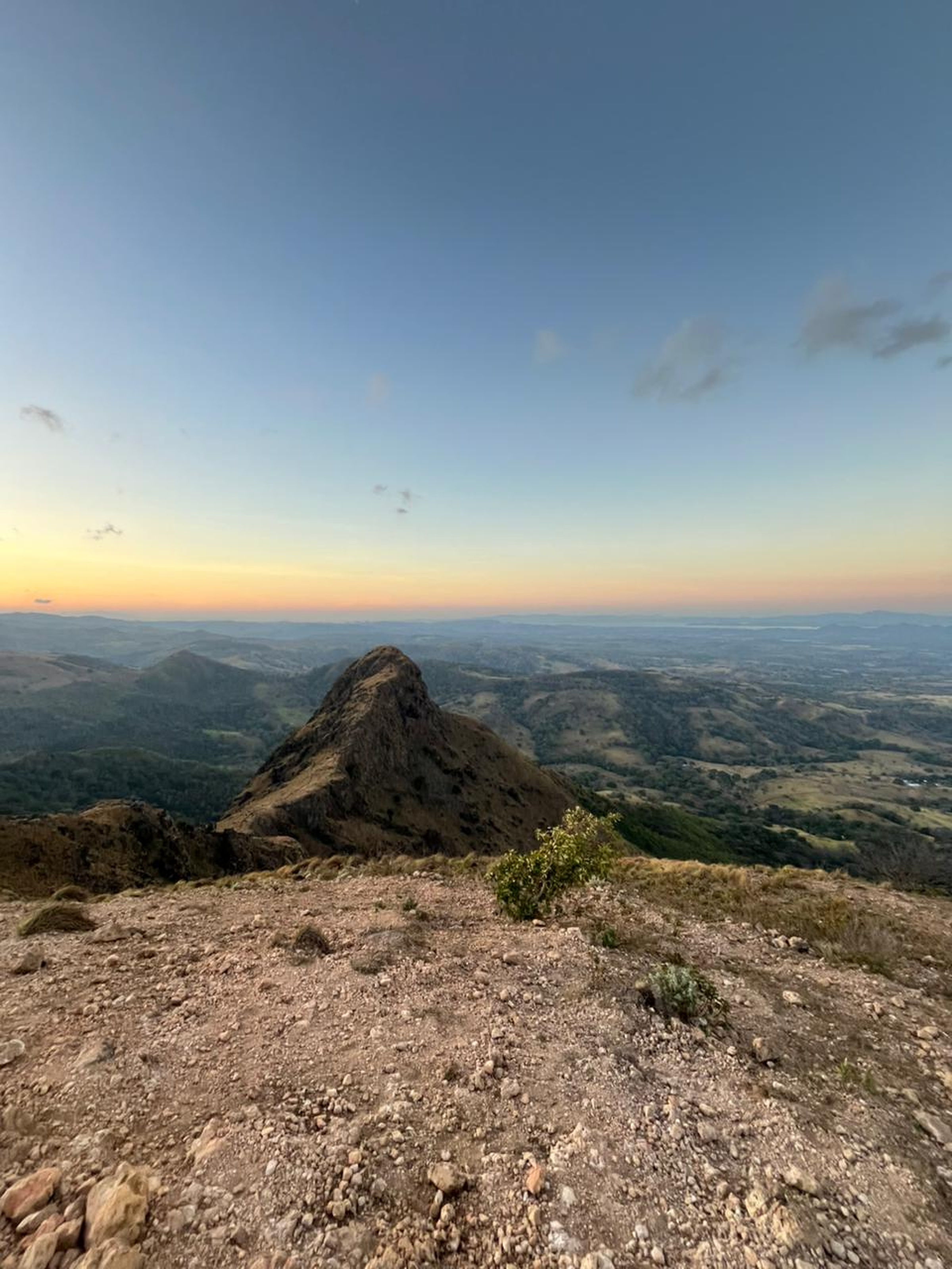 A dramatic landscape view from a mountain summit showing a rocky peak and a vast green valley at sunset