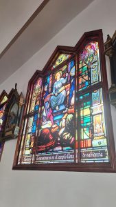 A stained glass window depicting a scene with a central figure seated, wearing a blue robe and a halo, gesturing with one hand. Surrounding the figure are various smaller characters, some in robes, appearing to listen or engage. The glass displays vibrant colors, including blues, reds, and yellows, with intricate details and patterns. The text at the bottom translates to "They found him in the temple full of wisdom." The window is framed in rich wood and is set against a light-colored wall.