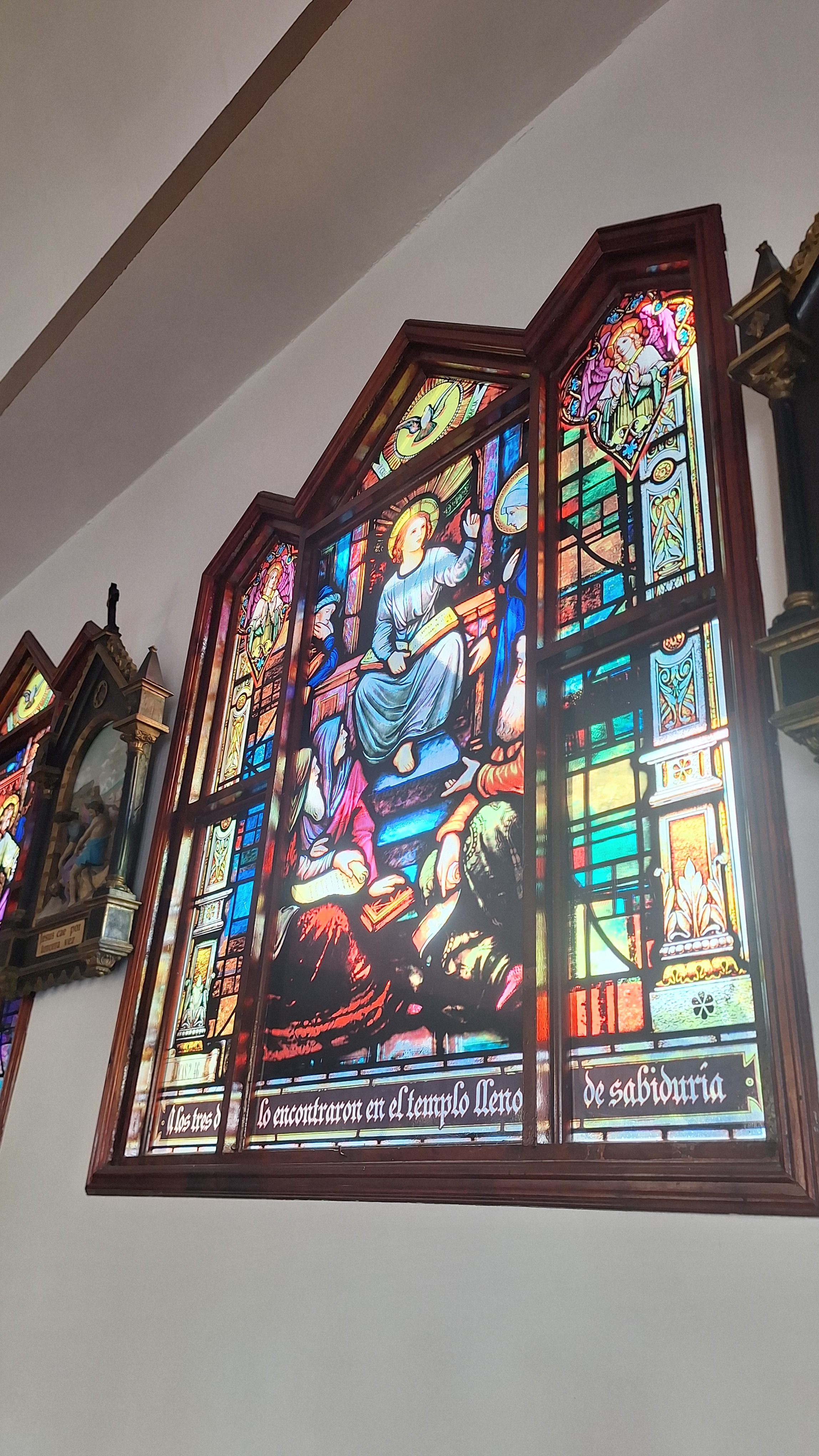 A stained glass window depicting a scene with a central figure seated, wearing a blue robe and a halo, gesturing with one hand. Surrounding the figure are various smaller characters, some in robes, appearing to listen or engage. The glass displays vibrant colors, including blues, reds, and yellows, with intricate details and patterns. The text at the bottom translates to "They found him in the temple full of wisdom." The window is framed in rich wood and is set against a light-colored wall.