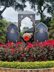 An ornate garden display with a decorative arch featuring Arabic calligraphy, a central traditional drum, and red flowers surrounded by greenery under a cloudy sky.
