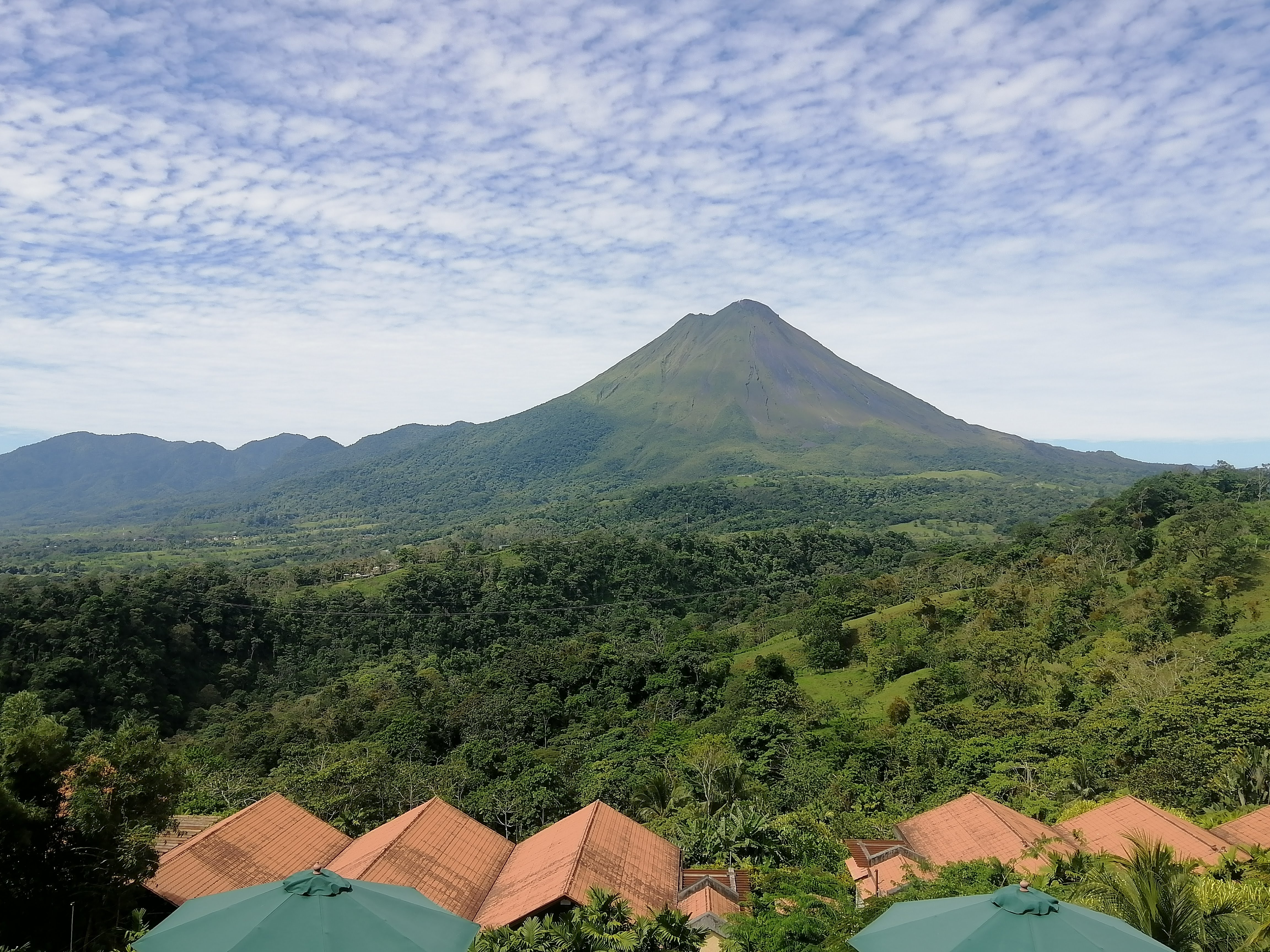 A scenic view of a volcano surrounded by lush green hills and forests. In the foreground, there are rooftops of buildings with terracotta-colored tiles and green umbrellas, leading to a vibrant landscape of trees and mountains under a partly cloudy sky. The volcano rises majestically in the background, providing a dramatic focal point in the picturesque setting.
