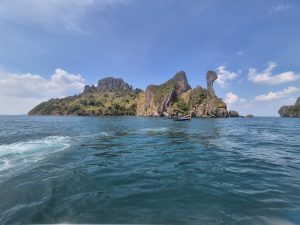Rocky island with steep cliffs rising from the sea under a clear blue sky, with a small boat floating on calm water in the foreground.
