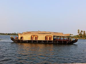 A traditional houseboat with a woven roof and wooden windows sails on calm blue water, surrounded by palm trees under a clear sky.