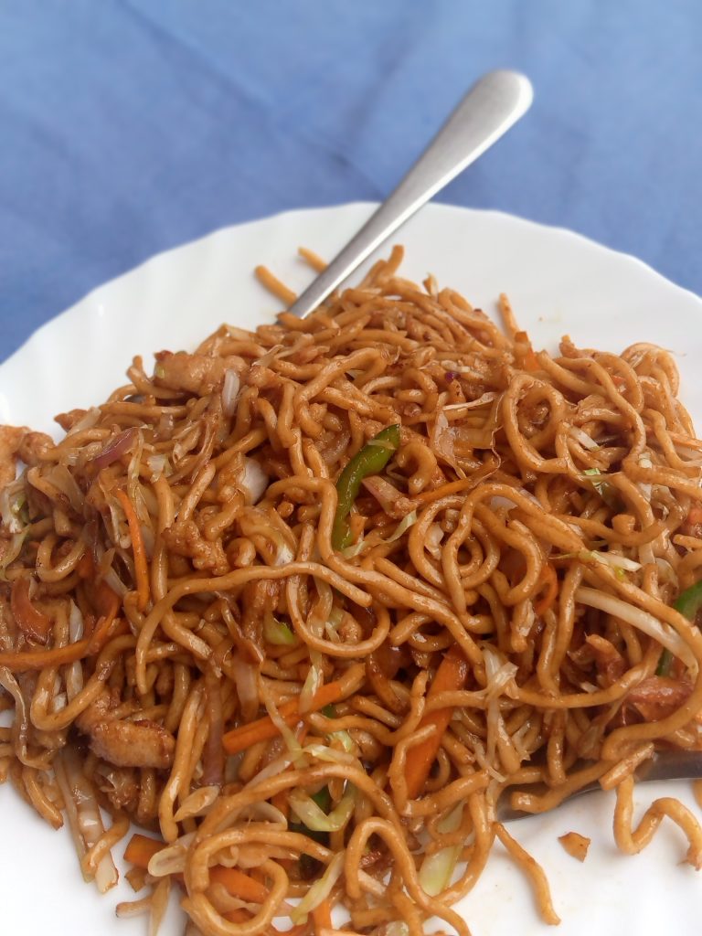 A plate of stir-fried noodles with meat and vegetables, with a fork on a blue background.