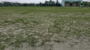 A wide view of a large open field with patches of green grass and sand under a cloudy sky.