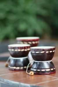 Three small, cylindrical drums are placed on a wooden surface. The drums are primarily black with a maroon finish and feature decorative elements, including a pattern of small, light-colored dots around their circumference. Each drum is shown resting on its wider base, and some strings attached to beads can be seen, adding to their decorative style. The background is blurred with greenery, emphasizing the drums in the foreground.

