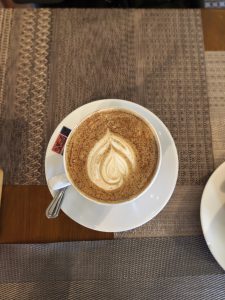 White ceramic cup with leaf-shaped latte art and cinnamon sprinkles, accompanied by a spoon, rests on a white saucer with a logo, placed on a textured, multi-patterned woven placemat.