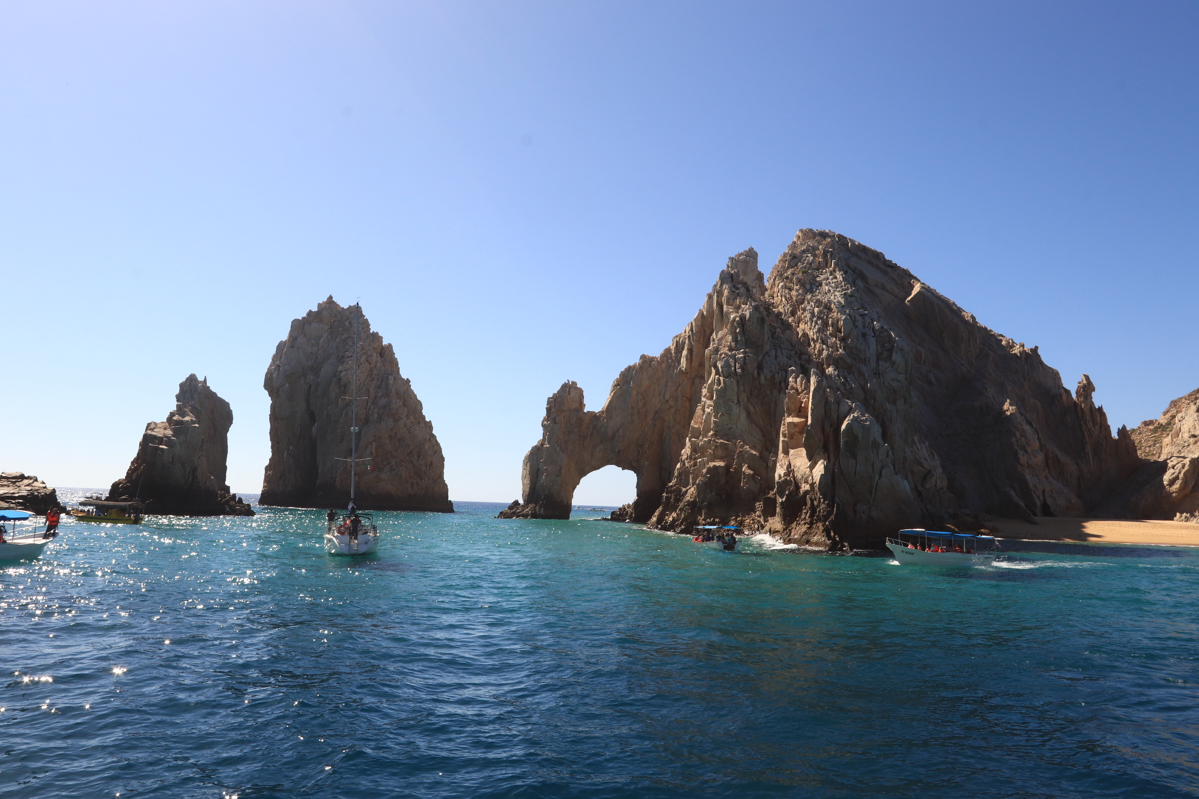 The Arch of Los Cabos stands between towering rock formations, with boats crossing the bright blue water beneath the clear coastal sky.