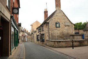 A narrow, winding street in a European town, lined with stone and brick buildings, including a historic structure with a tall chimney and storefronts with hanging signs.
