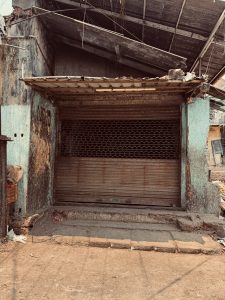 A storefront with a closed metal gate, partially obscured by a rusty, weathered awning. The building shows signs of wear, with peeling paint and exposed bricks.