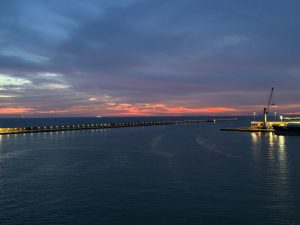 This image shows a calm sunset over the ocean near a harbor. The sky is painted with soft shades of pink, orange, and blue as the sun sets on the horizon. A long pier stretches into the sea, lined with lights that reflect gently on the water. On one side, there are port structures and a crane, adding a contrast between nature and human activity. Th