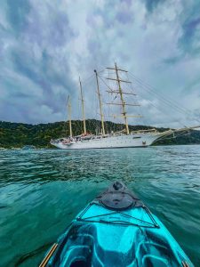 Vista desde un kayak en el agua hacia un velero blanco con mástiles altos. El mar es claro y tranquilo, rodeado de montañas verdes bajo un cielo nublado.