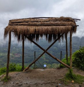 A rustic structure with a thatched roof made of grass sits atop wooden poles, overlooking a misty mountainous landscape.