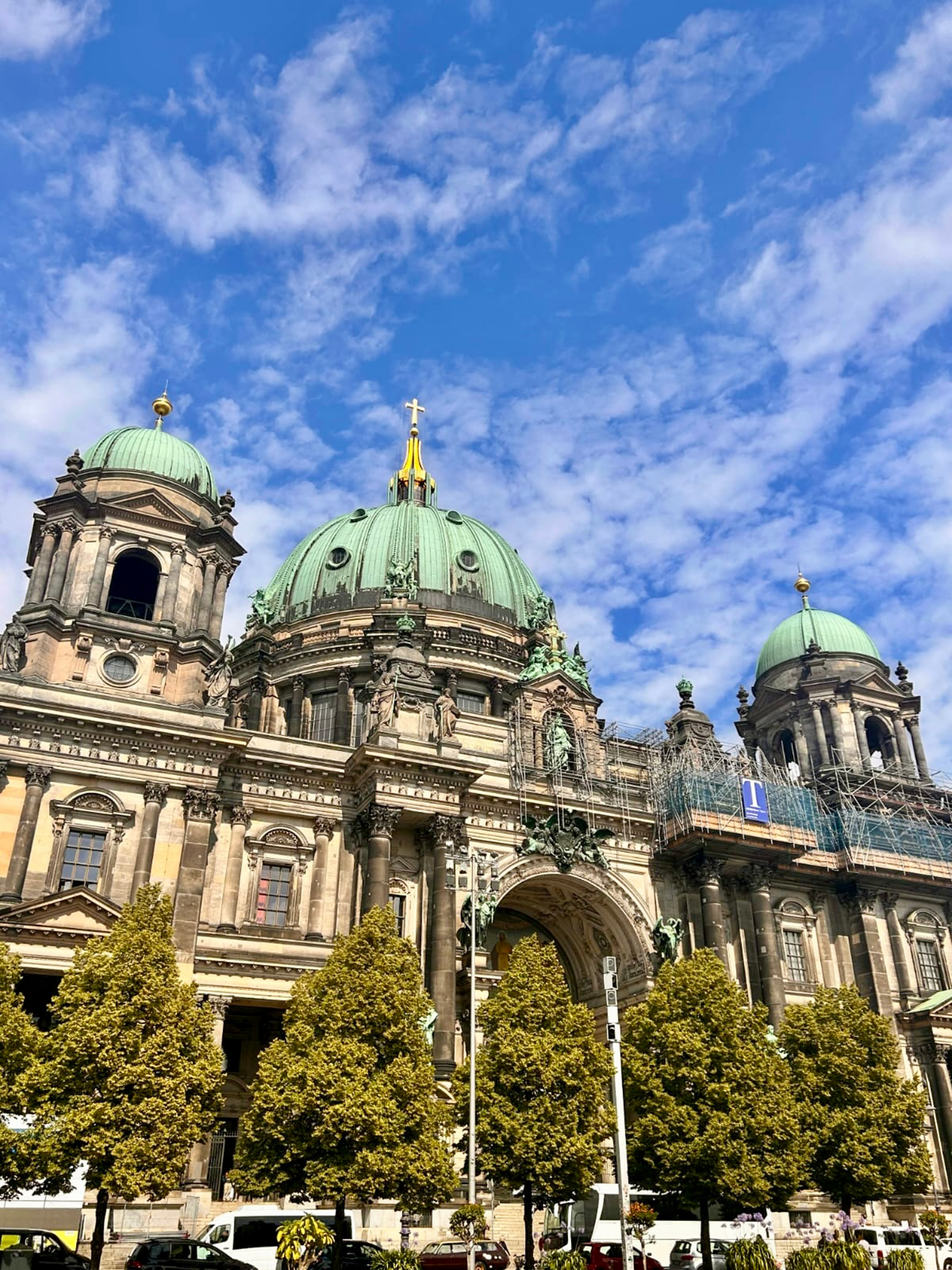 The image showcases a grand building with green domes and intricate architectural details, likely a historical or religious structure. The foreground features lush, vibrant trees, while the background displays a bright blue sky with scattered clouds. Scaffolding is visible on one side of the building, suggesting ongoing restoration or maintenance work.