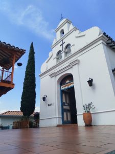 A white church with a bell tower and a cross on top, surrounded by a blue sky. In front, there is a tiled walkway leading to the door, which is framed by two lanterns. To the left, a tall, narrow evergreen tree rises next to the church, and there is a decorative pot with a plant at the base of the entrance. In the background, traditional architecture is visible, including a wooden balcony.
