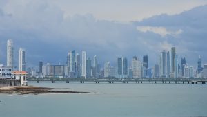 Panama City skyline with numerous high-rise buildings overlooking a calm body of water, featuring a long pier and a cloudy sky, viewed from a waterfront location.
