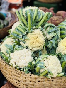 A woven basket filled with fresh cauliflowers, showcasing their creamy white heads and vibrant green leaves and stalks. The cauliflowers are piled high and vary in size, with some partially covered by leaves. In the background, hints of other produce can be seen, creating a colorful market scene.
