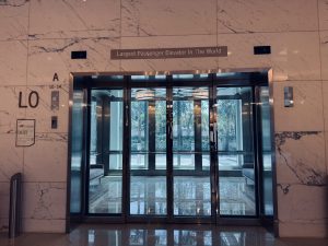 A large glass elevator in a building lobby with marble walls and a sign above it.